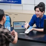 Bank teller in blue uniform hands a brochure to a male customer at a service desk with a laptop between them, in a bright office.