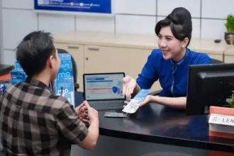 Bank teller in blue uniform hands a brochure to a male customer at a service desk with a laptop between them, in a bright office.