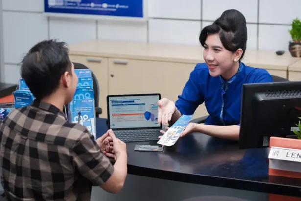 Bank teller in blue uniform hands a brochure to a male customer at a service desk with a laptop between them, in a bright office.