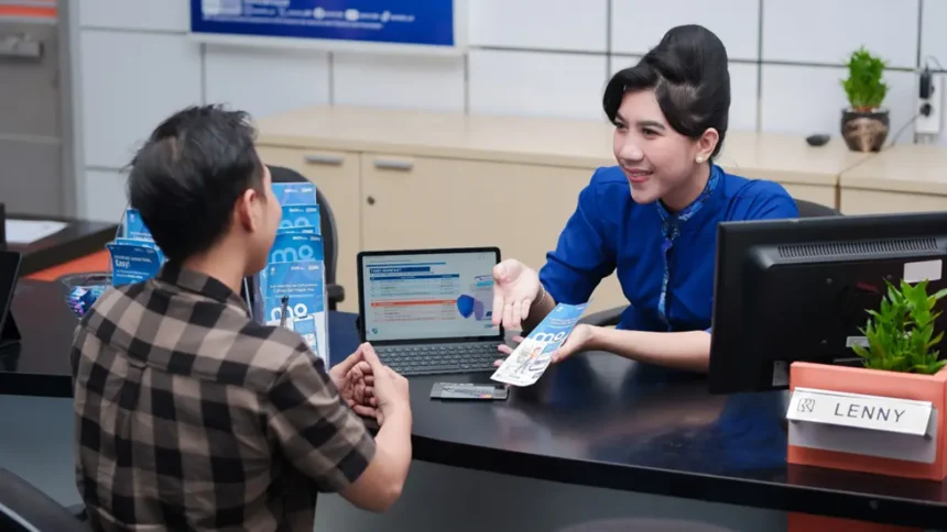 Bank teller in blue uniform hands a brochure to a male customer at a service desk with a laptop between them, in a bright office.