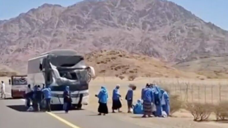 Damaged bus on a desert highway with a group of people in blue clothing nearby; rocky mountains in the background.