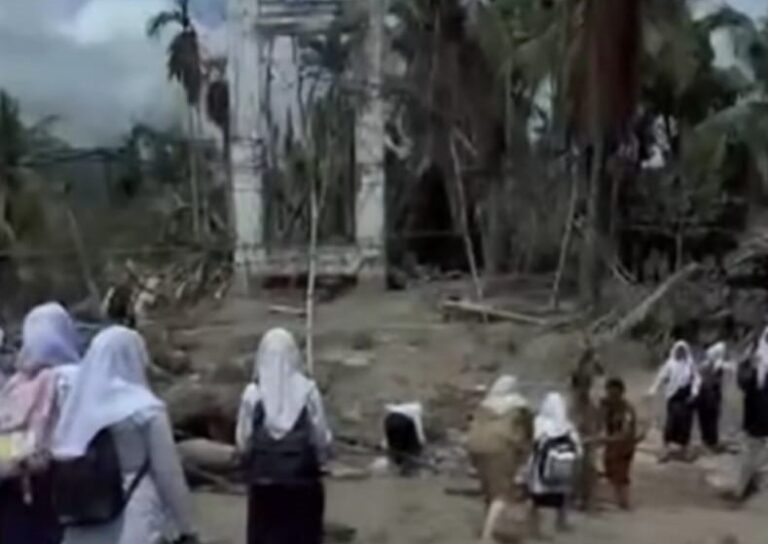 Group of people in headscarves walking through a ruined village with collapsed structures and debris in the background.