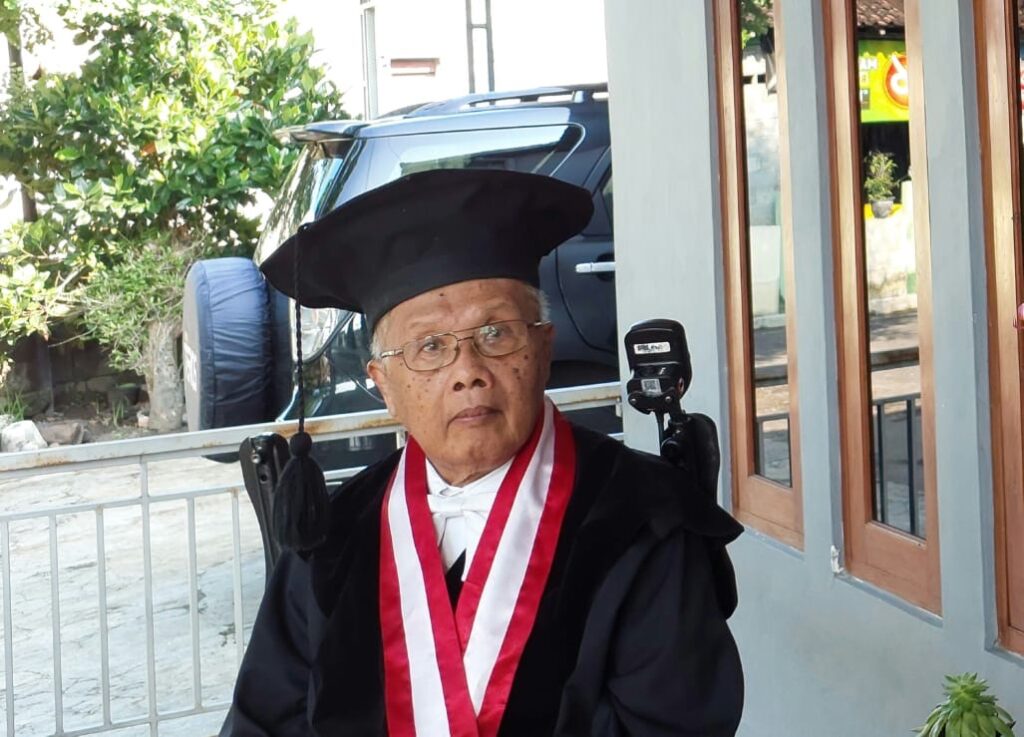 Elderly man in a graduation gown and cap with a red-and-white stole, seated in a wheelchair outside a building.