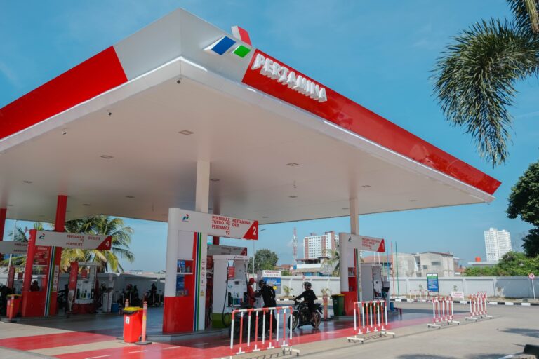 Pertamina gas station with a red-and-white canopy, fueling pumps, and motorcyclists charging at the pumps under a blue sky.