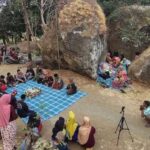 People gathering around a blue checkered tarp for a communal meal in a rocky, tree-filled outdoor area.