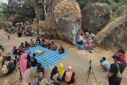 People gathering around a blue checkered tarp for a communal meal in a rocky, tree-filled outdoor area.