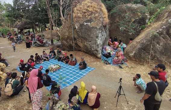 People gathering around a blue checkered tarp for a communal meal in a rocky, tree-filled outdoor area.