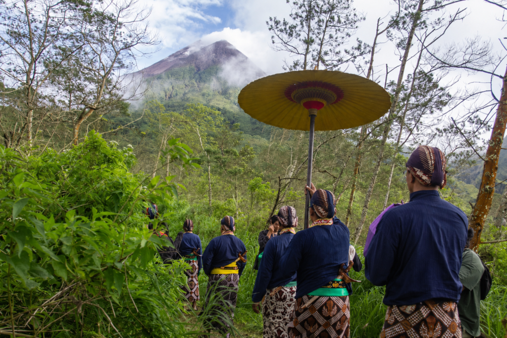 Group of people in traditional sarongs stand in a green forest; a person holds a large yellow parasol with a misty mountain in the background.