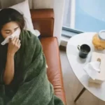 Woman wrapped in a green blanket on a couch, blowing her nose with a tissue. Table with tea, fruit, and tissues nearby; sick at home.