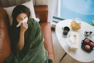 Woman wrapped in a green blanket on a couch, blowing her nose with a tissue. Table with tea, fruit, and tissues nearby; sick at home.