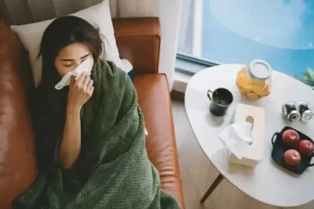 Woman wrapped in a green blanket on a couch, blowing her nose with a tissue. Table with tea, fruit, and tissues nearby; sick at home.