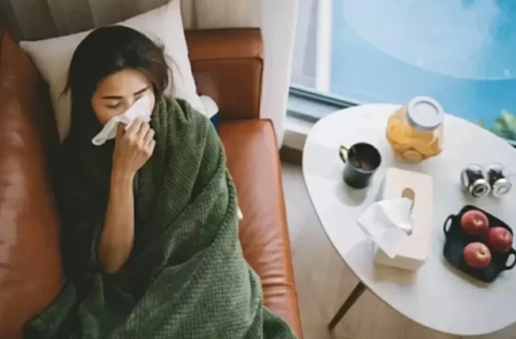 Woman wrapped in a green blanket on a couch, blowing her nose with a tissue. Table with tea, fruit, and tissues nearby; sick at home.