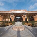 Symmetrical courtyard with curved wooden arches, benches, and a circular fountain in front of a building with Arabic signage under a blue sky.