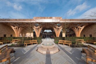 Symmetrical courtyard with curved wooden arches, benches, and a circular fountain in front of a building with Arabic signage under a blue sky.