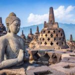 Stone Buddha statue in meditation at Borobudur, surrounded by bell-shaped stupas and stone terraces against a blue sky and distant hills