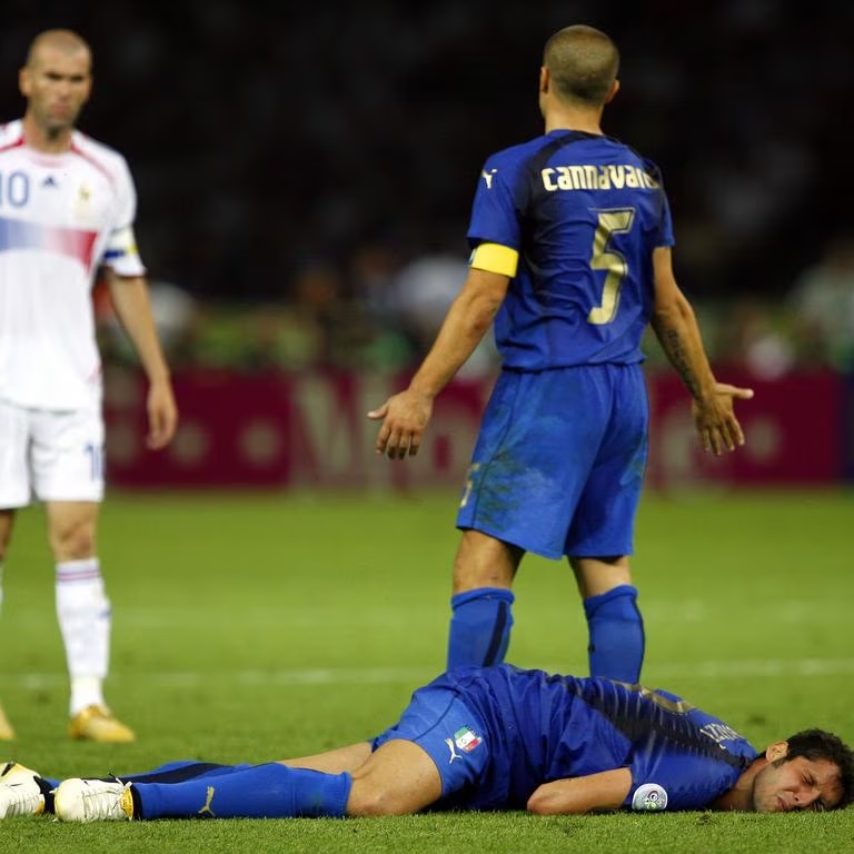 cannavaro asking Zidane what happens by News Corp Australia - Mabur.co Soccer scene: a blue‑kit player lies on the pitch after a fall, with another blue player standing nearby and a white‑kit opponent watching in the background.