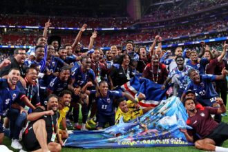 Soccer team celebrates on the field, arms raised, with banners and flags as the stadium crowds cheer in the background.
