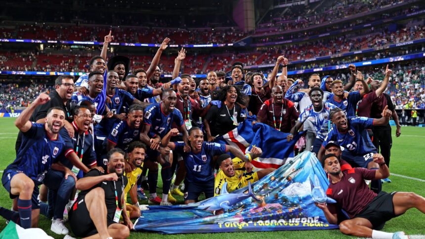 Soccer team celebrates on the field, arms raised, with banners and flags as the stadium crowds cheer in the background.
