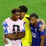 Three soccer players embrace on a green pitch, wearing pink, yellow, and blue jerseys.