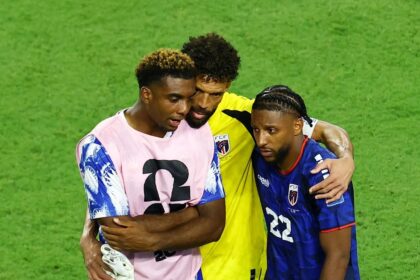 Three soccer players embrace on a green pitch, wearing pink, yellow, and blue jerseys.