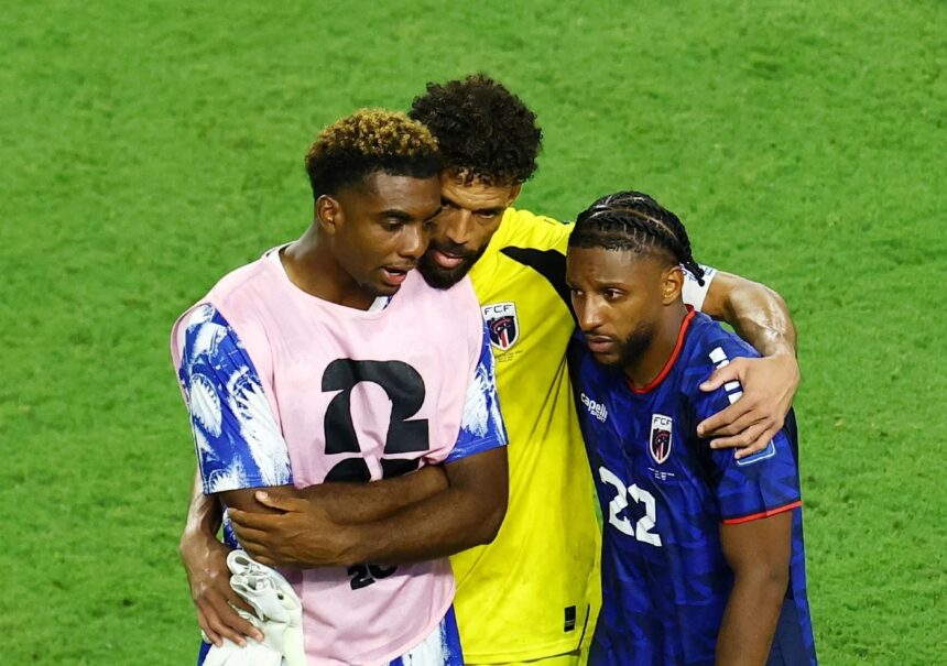 Three soccer players embrace on a green pitch, wearing pink, yellow, and blue jerseys.