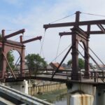 Rusty steel bridge framework under construction over a canal, with palm trees and a distant city skyline in view.