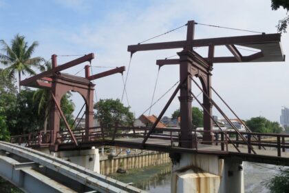 Rusty steel bridge framework under construction over a canal, with palm trees and a distant city skyline in view.