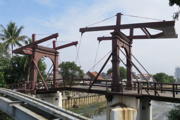 Rusty steel bridge framework under construction over a canal, with palm trees and a distant city skyline in view.