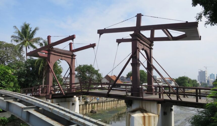 caption - Mabur.co Rusty steel bridge framework under construction over a canal, with palm trees and a distant city skyline in view.