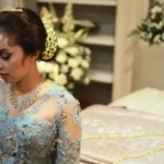 Young woman in a light blue, heavily embroidered gown with a tiara and necklace, looking down in a wedding or formal setting.