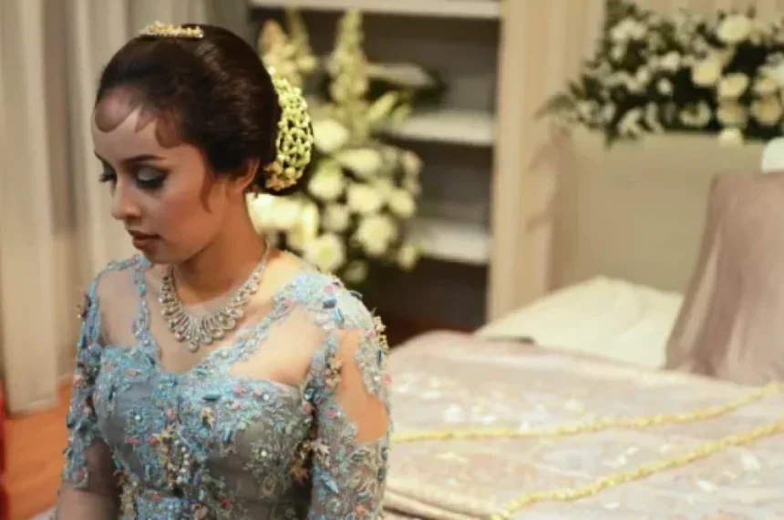 Young woman in a light blue, heavily embroidered gown with a tiara and necklace, looking down in a wedding or formal setting.