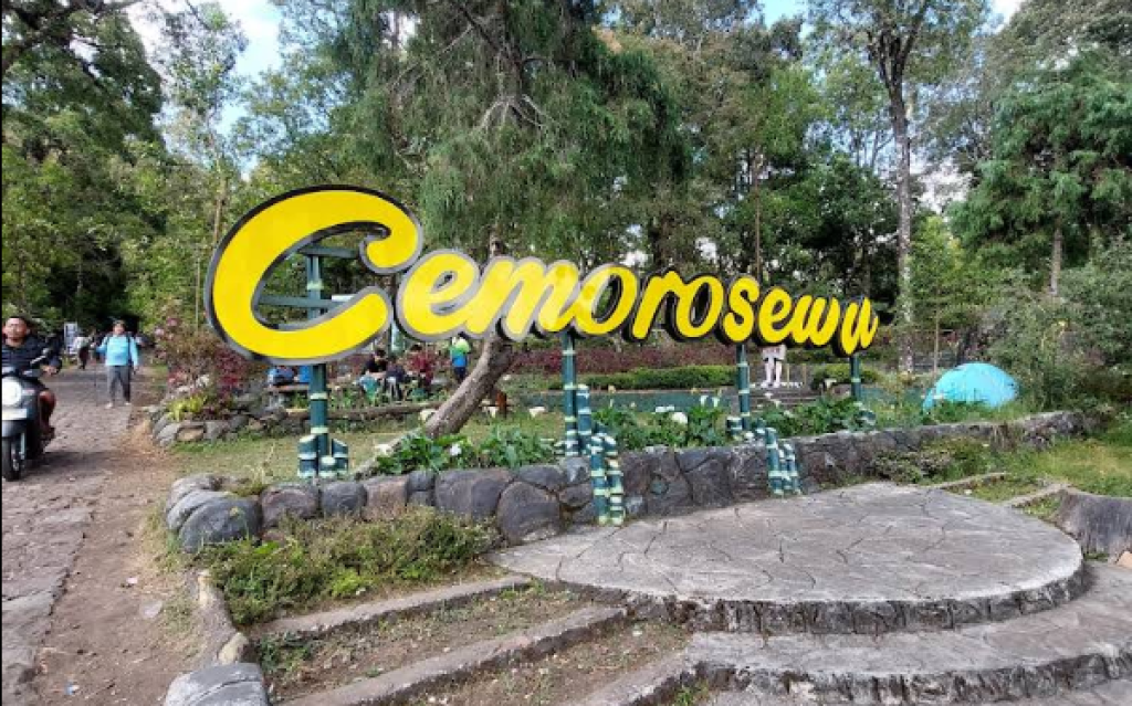 Large yellow cursive sign reading 'Gemorosew' in a park with a circular stone platform and greenery behind it.