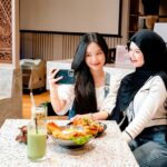Two women sit at a spotted table, one in a hijab, taking a selfie with a phone as they share a meal.