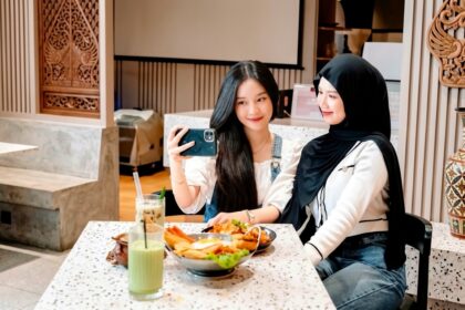 Two women sit at a spotted table, one in a hijab, taking a selfie with a phone as they share a meal.