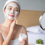 Woman applies a white facial mask while smiling, standing in a bedroom with a mirror nearby.