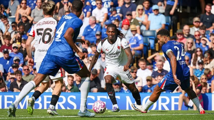 Soccer players in blue and white compete for the ball on a sunny stadium pitch with a crowd watching behind them, during a match.