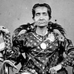 Portrait of a person seated in an ornate chair, wearing ceremonial, embellished clothing with large medals and jewelry in a black-and-white photo.