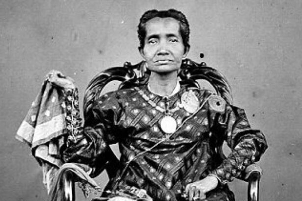 Portrait of a person seated in an ornate chair, wearing ceremonial, embellished clothing with large medals and jewelry in a black-and-white photo.