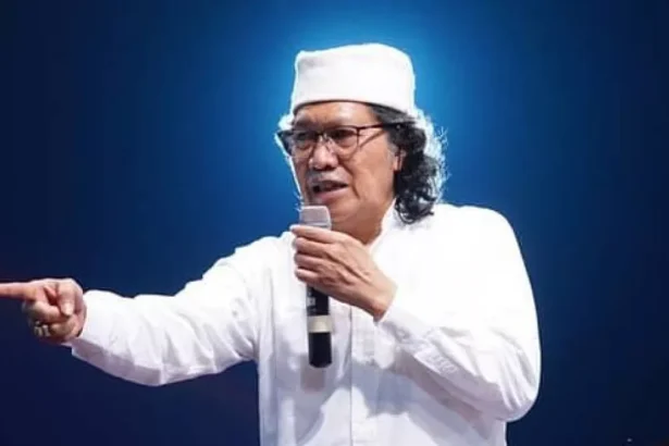Man on stage wearing a white outfit and hat, holding a microphone and pointing toward the audience.