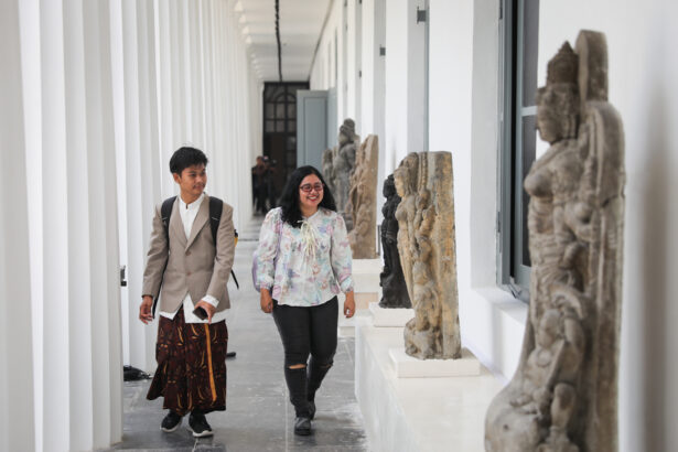 Two travelers walk down a bright hallway of a museum, examining stone statues on display along the wall.