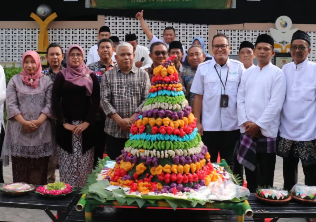 Group of people in traditional attire posing beside a tall colorful tiered fruit centerpiece (tumpeng) at a celebration.