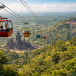 Red cable car with passengers gliding above a lush valley toward Angkor temples in the distance.