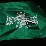 Green flag on a wooden pole with white Arabic script and a globe emblem against a black background, fluttering in the wind.