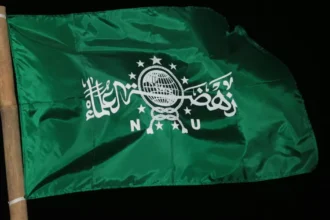 Green flag on a wooden pole with white Arabic script and a globe emblem against a black background, fluttering in the wind.