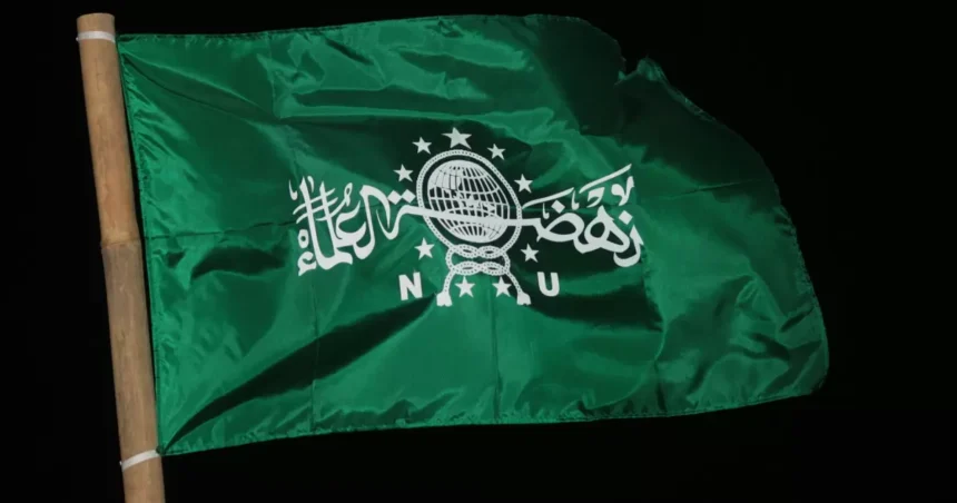 Green flag on a wooden pole with white Arabic script and a globe emblem against a black background, fluttering in the wind.