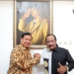 Two men shake hands in front of a large framed painting on a white wall behind them.