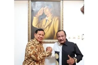 Two men shake hands in front of a large framed painting on a white wall behind them.