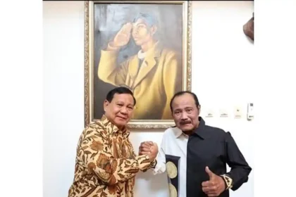 Two men shake hands in front of a large framed painting on a white wall behind them.