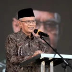 Smiling man wearing a batik shirt and black cap speaks at a podium with a microphone.