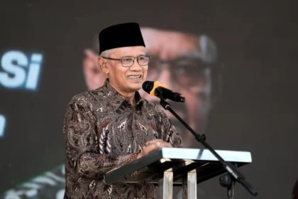 Smiling man wearing a batik shirt and black cap speaks at a podium with a microphone.